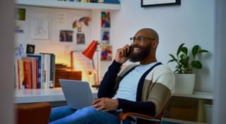 A man in his office, he is on the phone and has his laptop on his lap.